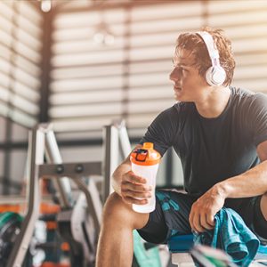 Portait of athletic man in headphones looking aside while listening to music and holding a towel and a classic fitness shaker with pre-workout drink in it. Horizontal shot