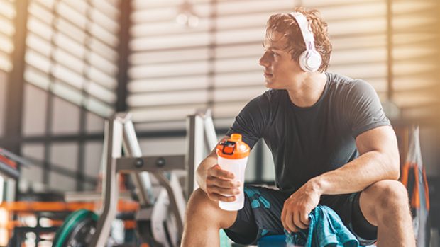 Portait of athletic man in headphones looking aside while listening to music and holding a towel and a classic fitness shaker with pre-workout drink in it. Horizontal shot